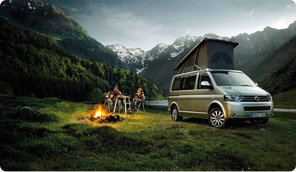 A VW campervan parked infront of some mountains with 2 people sitting infront of a campfire.