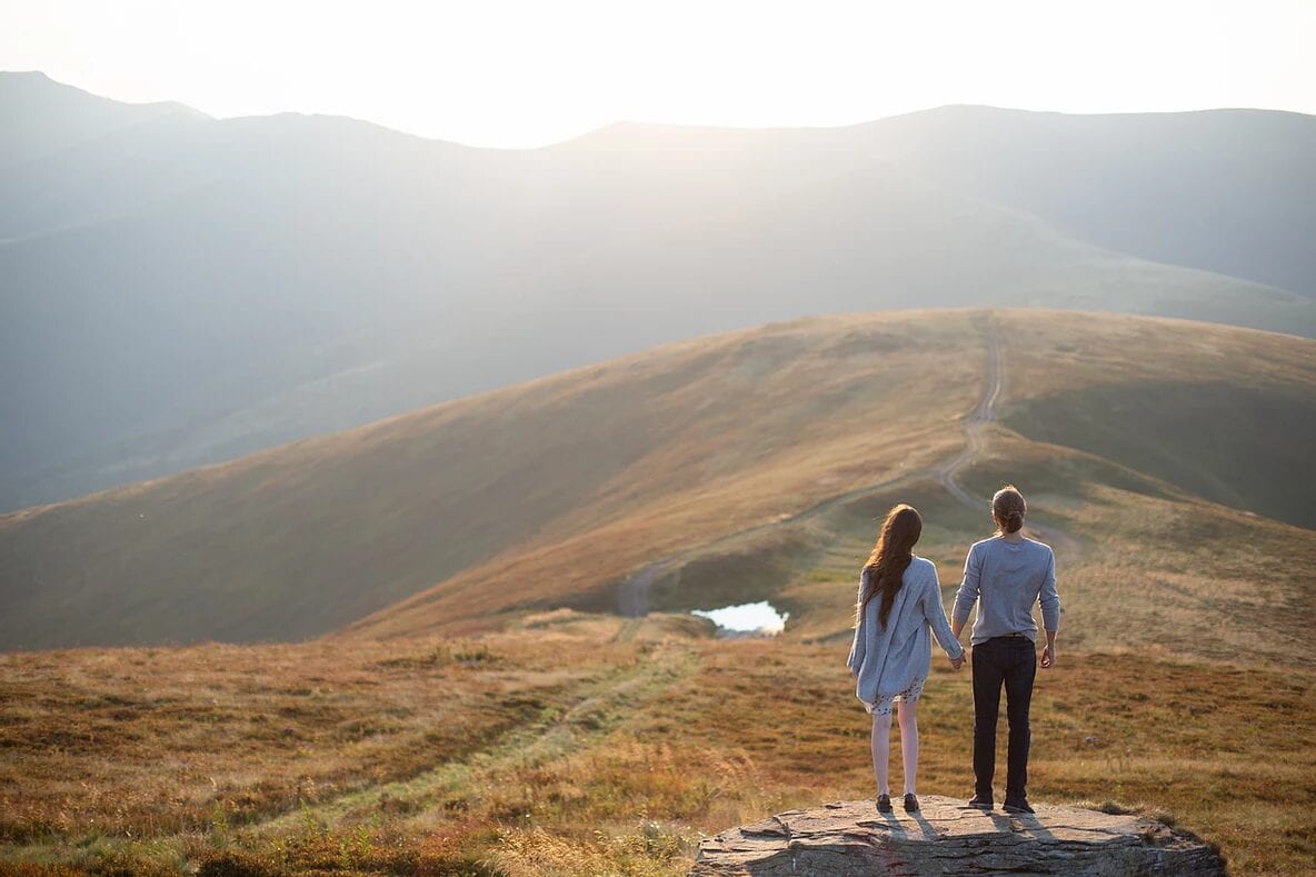 Two people holding hands at the top of a hill and mountain range.