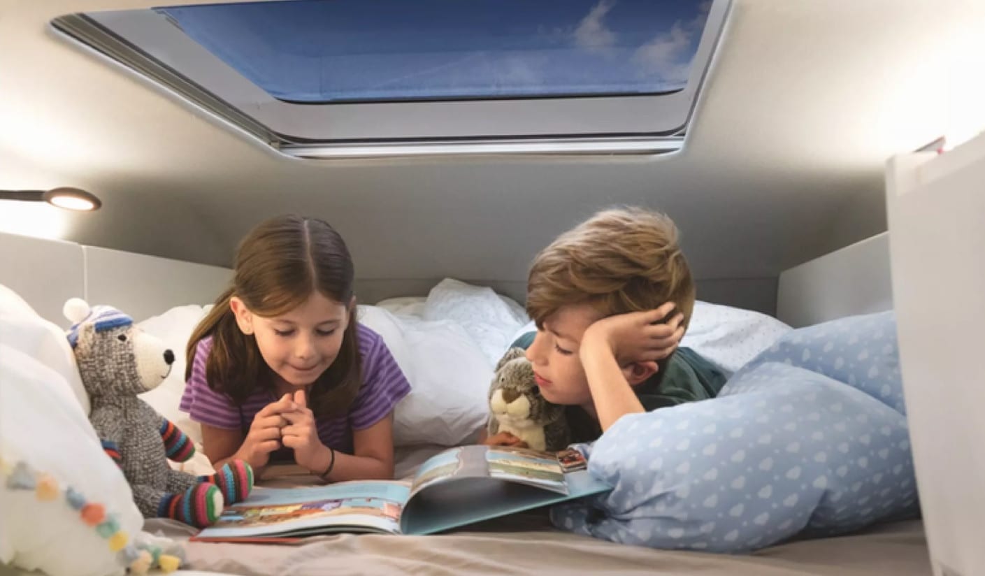 2 children led on a bed inside of a VW Grand California reading a magazine together.